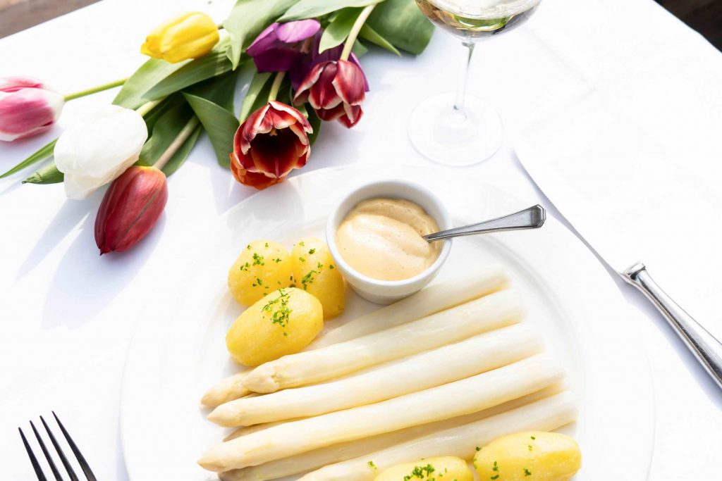 Festlich gedeckter Tisch mit weißem Spargel, Sauce, Kartoffeln und frischen Tulpen. Die helle Tischdekoration wirkt elegant und saisonal.