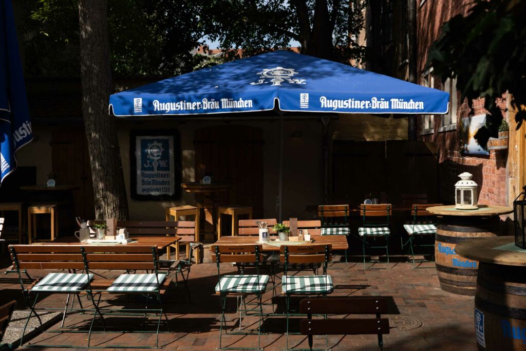 Gemütlicher Innenhof-Biergarten mit langen Holztischen, Bierbänken und einem großen blauen Augustiner-Bräu-Sonnenschirm vor einer Backsteinfassade.