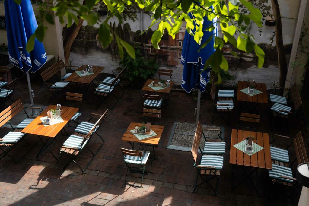 Blick von oben auf einen schattigen Innenhof-Biergarten mit Holztischen, Stühlen und blauen Sonnenschirmen zwischen Backsteinfassade und grünen Pflanzen.