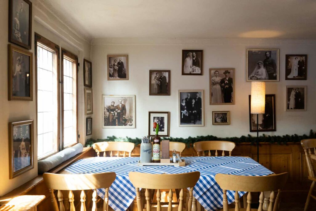 Gemütlicher Gastraum mit Holztischen, karierten Tischdecken, großen Fenstern und einer Bilderwand mit historischen Familienfotos.