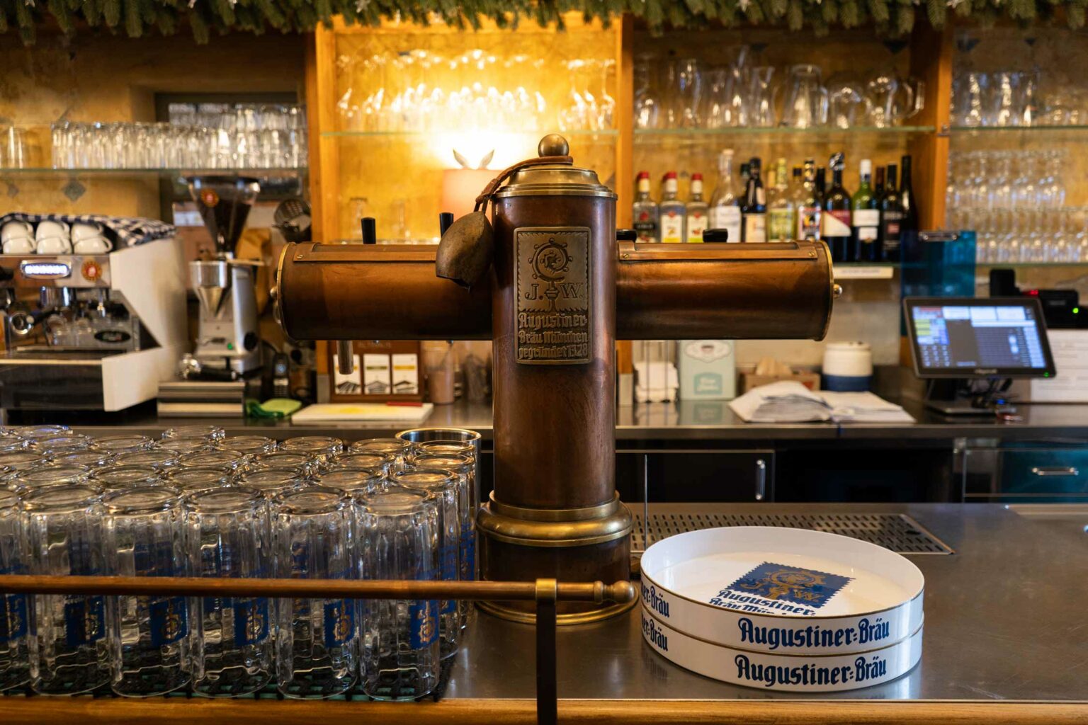 Traditioneller Augustiner-Zapfhahn an der Bar mit aufgereihten Biergläsern im Vordergrund. Kupferdetails und warmes Licht betonen die rustikale Wirtshausatmosphäre.