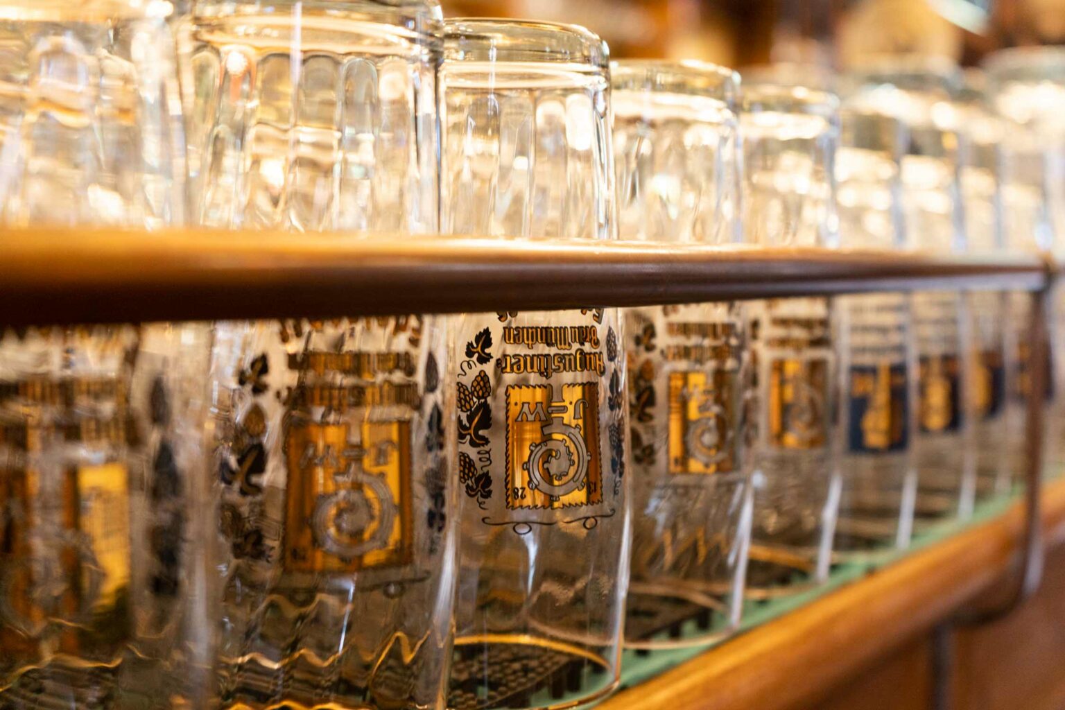 Nahaufnahme mehrerer Augustiner-Biergläser auf einem Regal hinter der Bar. Warme Lichtreflexe und goldene Wappen sorgen für eine stimmungsvolle Brauhausatmosphäre.