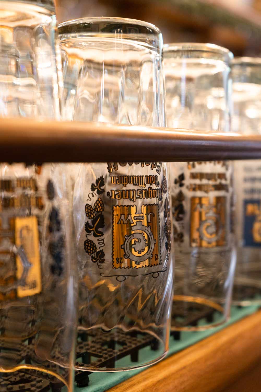 Nahaufnahme umgedrehter Augustiner-Biergläser auf einem Regal hinter der Bar. Die enge Perspektive und warmen Lichtreflexe sorgen für eine authentische Brauhausatmosphäre.