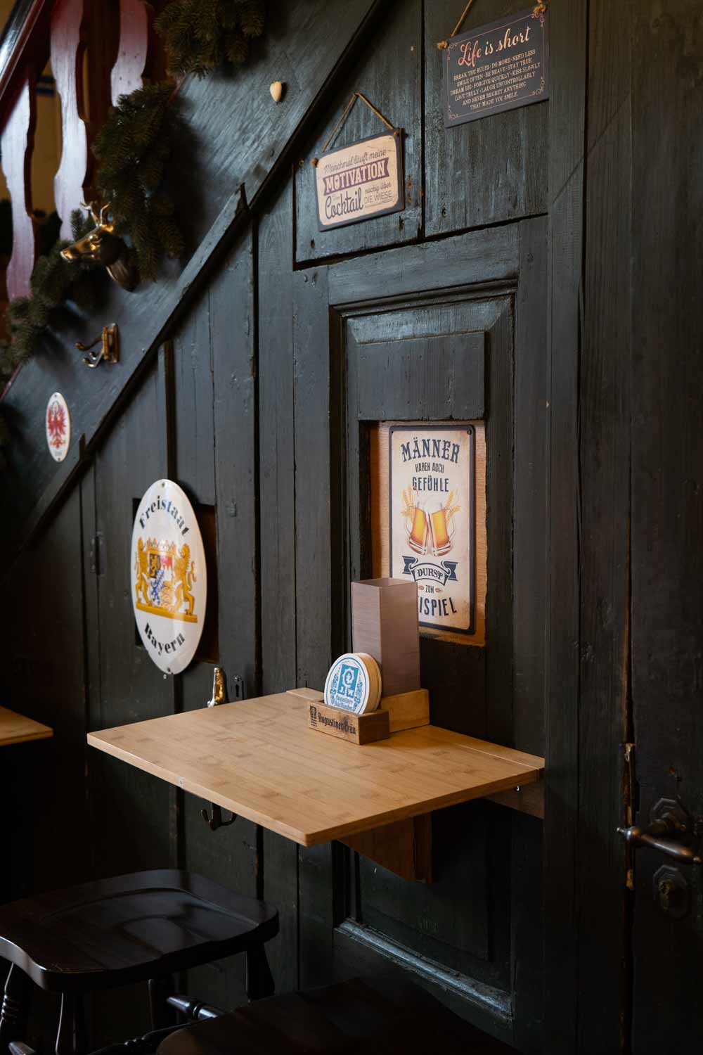 Kleiner Stehtisch vor dunkler Holzverkleidung mit historischen Bier- und Wirtshausschildern. Die rustikale Dekoration vermittelt eine traditionelle, gemütliche Augustiner-Atmosphäre.