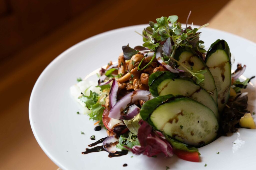 Nahaufnahme eines frischen Salats mit Gurken, roten Zwiebeln, Kräutern und weiteren Zutaten auf einem weißen Teller. Die natürliche Anrichtung und das warme Licht lassen das Gericht frisch und appetitlich wirken.