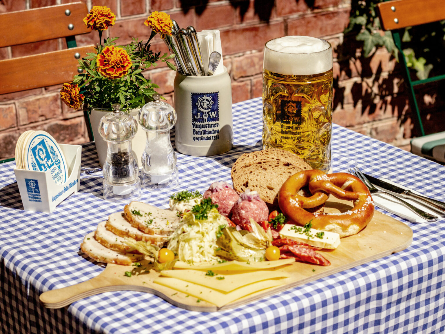 Deftig gedeckter Wirtshaustisch mit Bier, Brezn, Fleisch, Käse und weiteren bayerischen Spezialitäten. Blau-weiß karierte Tischdecke und rustikale Details unterstreichen die traditionelle Atmosphäre.