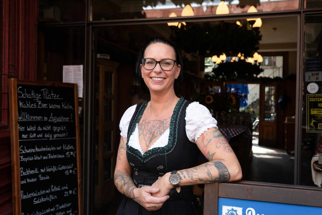 Freundlich lächelnde Mitarbeiterin in traditioneller Kleidung vor dem Eingang eines Augustiner-Restaurants. Im Hintergrund sind der warm beleuchtete Gastraum, eine Speisekarte auf einer Tafel und rustikale Wirtshausdetails zu sehen.