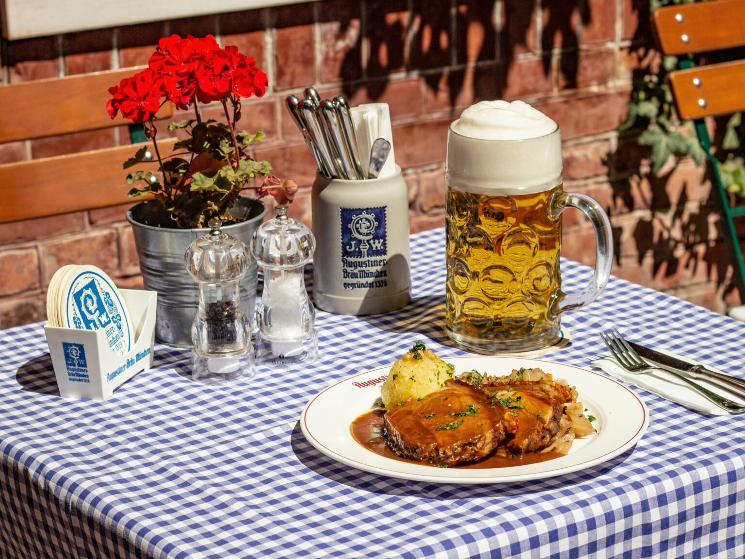 Traditionelles Wirtshausgericht mit Bierkrug auf einem blau-weiß gedeckten Tisch. Blumen, Gewürzmühlen und rustikale Details schaffen eine gemütliche Biergartenstimmung.