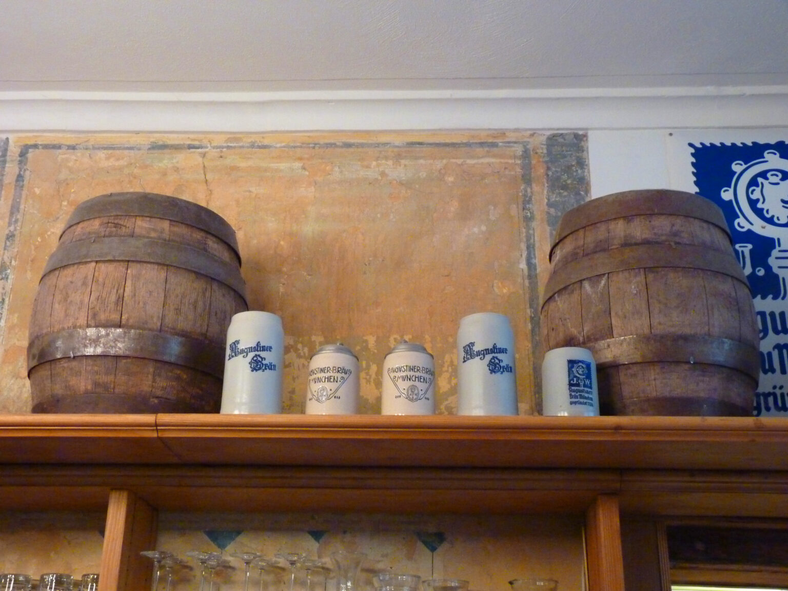 Rustikale Wanddekoration mit alten Holzfässern und verschiedenen Augustiner-Bierkrügen auf einem Holzregal. Die warme Farbgebung und traditionelle Gestaltung vermitteln eine gemütliche Wirtshausatmosphäre.