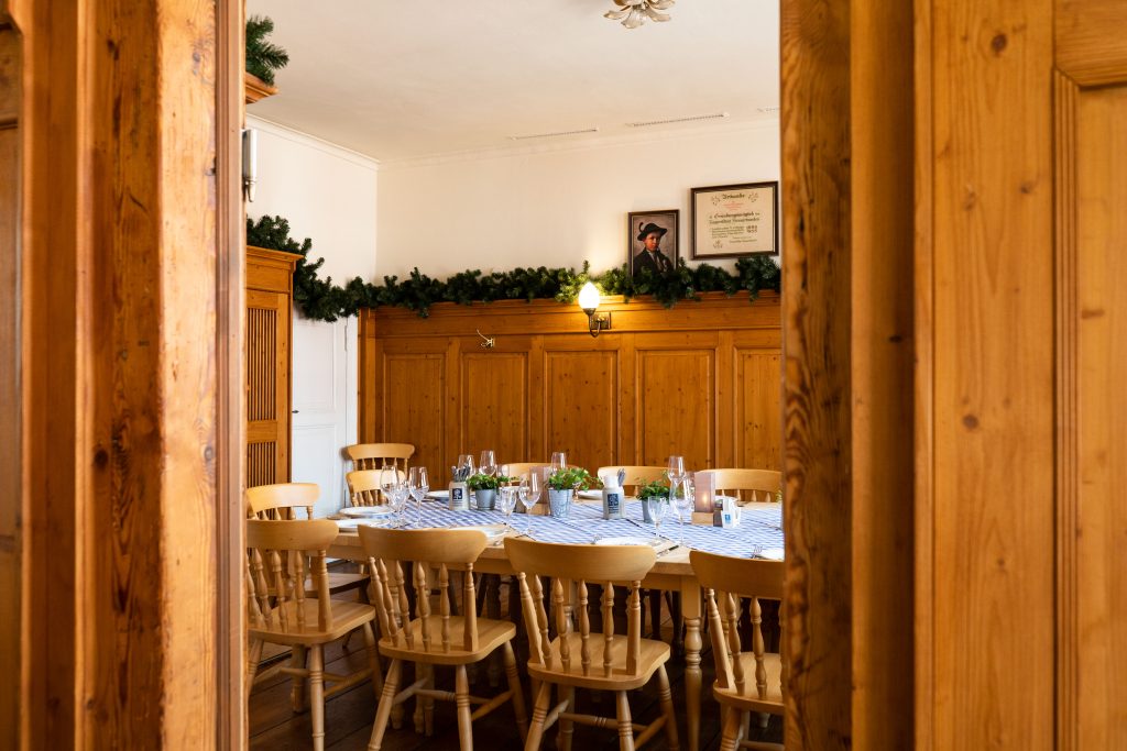 Blick durch eine Holztür in einen traditionell eingerichteten Gastraum mit langer festlich gedeckter Tafel, Holzstühlen und blau-weiß karierter Tischdecke. Warme Wandbeleuchtung, Holzvertäfelungen und grüne Dekoration schaffen eine gemütliche Atmosphäre für gesellige Anlässe.