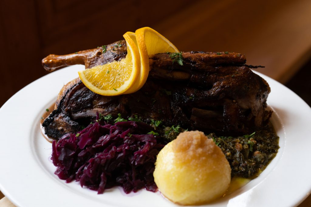 Traditionelles Wirtshausgericht mit knuspriger Entenkeule, Rotkohl, Knödel und Orangenscheibe auf einem weißen Teller. Warmes Licht und rustikale Anrichtung betonen den herzhaften, festlichen Charakter der Speise.
