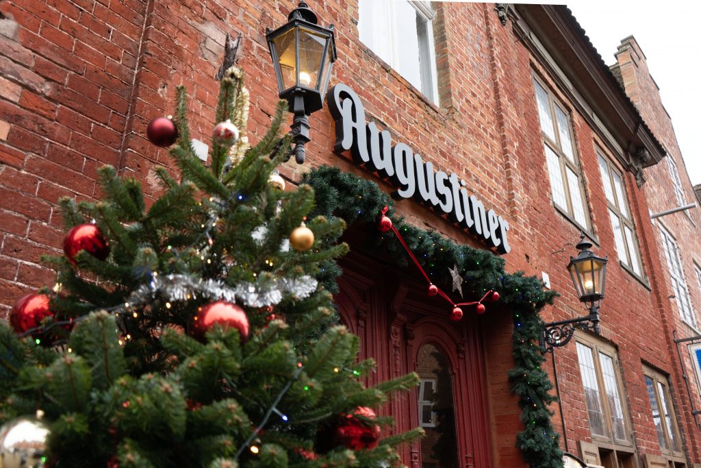 Weihnachtlich dekorierter Restaurant-Eingang mit roter Backsteinfassade, Augustiner-Schriftzug und geschmücktem Tannenbaum im Vordergrund. Laternen, grüne Girlanden und rote Kugeln schaffen eine festliche, einladende Atmosphäre.