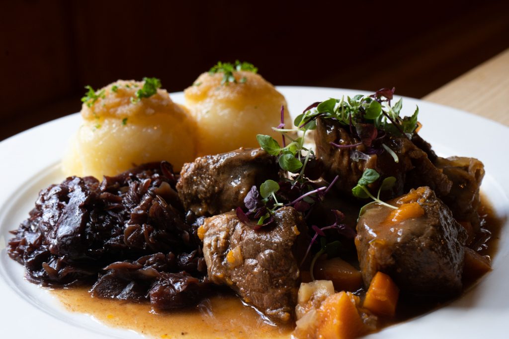 Herzhaftes Wirtshausgericht mit zartem Fleisch, dunkler Sauce, Rotkohl und zwei Knödeln auf einem weißen Teller. Frische Kräuter und warmes Licht setzen die traditionelle Speise appetitlich in Szene.