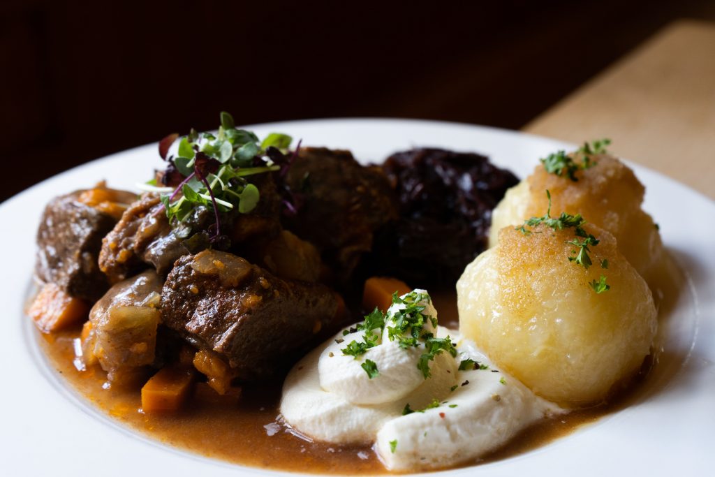 Nahaufnahme eines herzhaften Wirtshausgerichts mit Fleisch, Sauce, Knödeln und frischen Kräutern auf einem weißen Teller. Die warme Beleuchtung setzt das traditionelle Gericht stimmungsvoll in Szene.